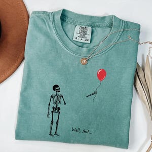 Pode incluir: Uma t-shirt verde s&aacute;lvia com um gr&aacute;fico de esqueleto preto, um bal&atilde;o vermelho flutuante segurado por uma m&atilde;o esquel&eacute;tica e o texto "Well, shit..." impresso em preto. A t-shirt tem uma etiqueta da marca Comfort Colors no decote interno.