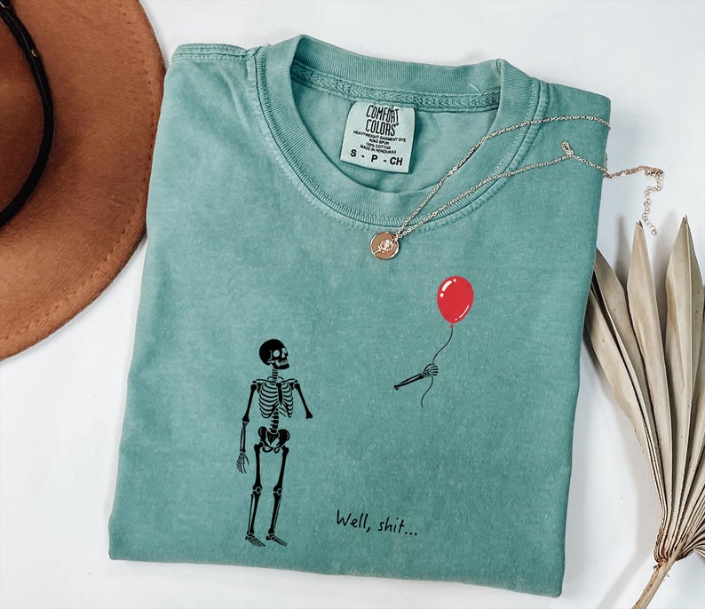 Pode incluir: Uma t-shirt verde s&aacute;lvia com um gr&aacute;fico de esqueleto preto, um bal&atilde;o vermelho flutuante segurado por uma m&atilde;o esquel&eacute;tica e o texto "Well, shit..." impresso em preto. A t-shirt tem uma etiqueta da marca Comfort Colors no decote interno.
