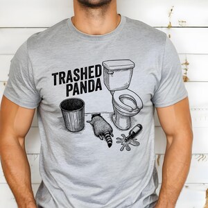 A line art illustration features a raccoon lying on its stomach on the floor, looking exhausted or passed out. Next to the animal is a spilled bottle with liquid pooling on the ground, a trash can, and a toilet. The text above reads TRASHED PANDA in bold, distressed letters.