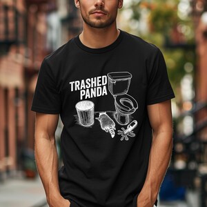 May include: Black t-shirt with a white graphic design. The design includes the text "Trashed Panda", a raccoon, a toilet, a trash can, and a spilled bottle. The shirt is short-sleeved.