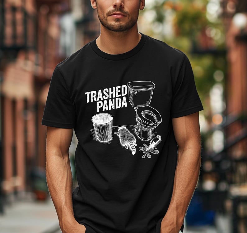 May include: Black t-shirt with a white graphic design. The design includes the text "Trashed Panda", a raccoon, a toilet, a trash can, and a spilled bottle. The shirt is short-sleeved.