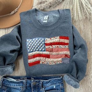 May include: A gray sweatshirt with a patchwork American flag design. The flag features white stars on a blue background and red and floral-patterned stripes. The sweatshirt is paired with blue jeans and a brown hat.