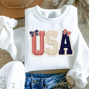 May include: White sweatshirt featuring the word "USA" in appliqued letters. The letters are decorated with red and white stripes, beige fabric, and navy blue polka dots. Each letter has a decorative bow.