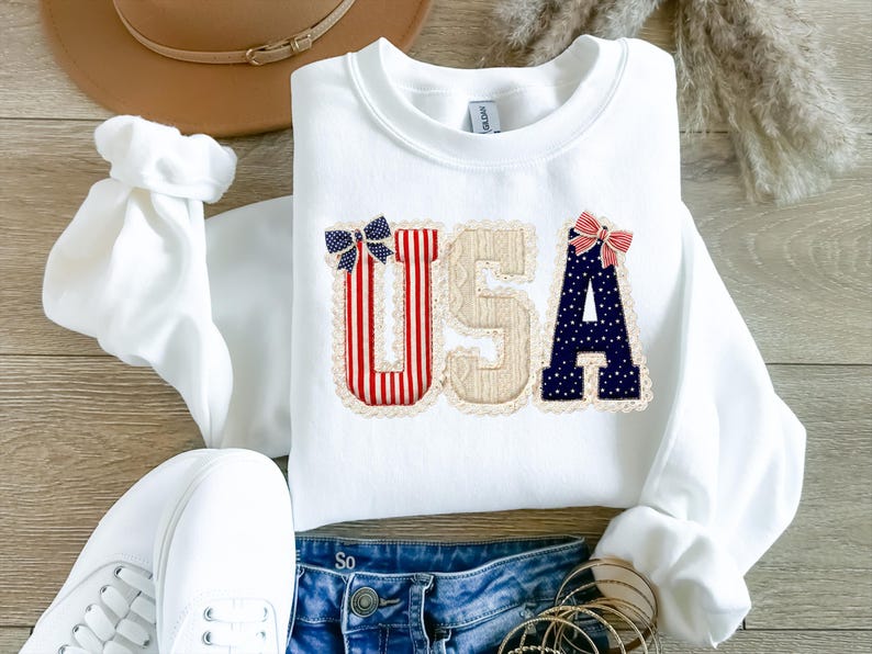 May include: White sweatshirt featuring the word "USA" in appliqued letters. The letters are decorated with red and white stripes, beige fabric, and navy blue polka dots. Each letter has a decorative bow.