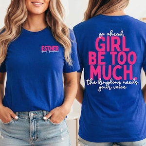 May include: A royal blue short-sleeve t-shirt featuring the text "ESTHER four:fourteen" in pink and white on the front chest. The back displays the pink text "go ahead, GIRL BE TOO MUCH." followed by white text "the kingdom needs your voice."