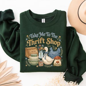 May include: Dark green sweatshirt with the text "Take Me To The Thrift Shop" and a graphic of a goose, chicken, bread box, bowls, and flowers. The sweatshirt has dark green cuffs and a white tag.