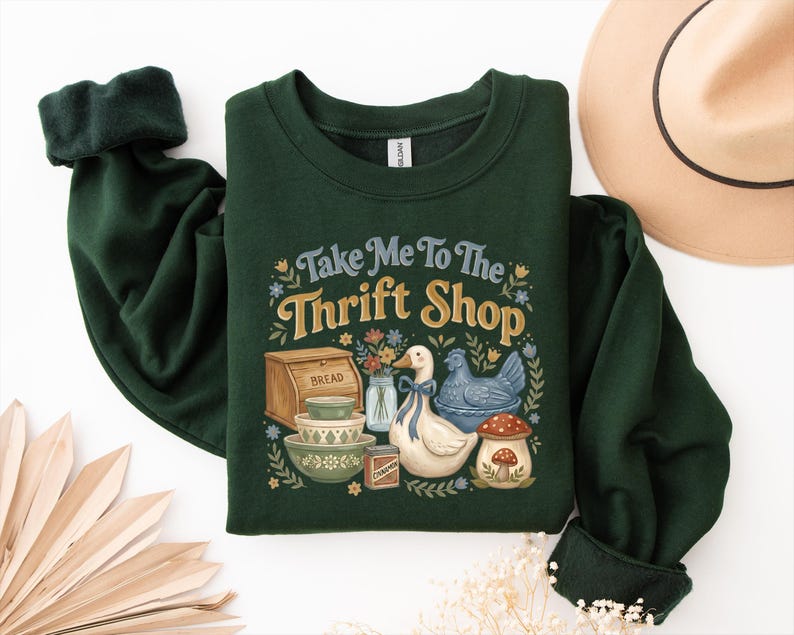 May include: Dark green sweatshirt with the text "Take Me To The Thrift Shop" and a graphic of a goose, chicken, bread box, bowls, and flowers. The sweatshirt has dark green cuffs and a white tag.