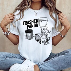May include: White t-shirt with a black graphic that reads "Trashed Panda." The graphic depicts a trash can, a toilet, a raccoon, and a spilled bottle. The design is a humorous illustration.