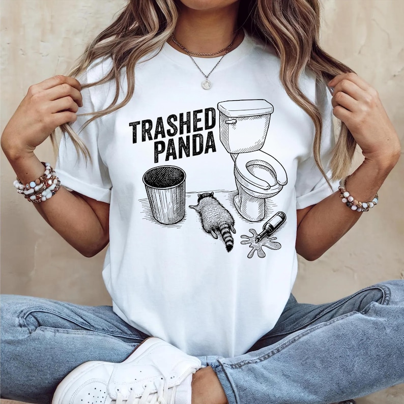May include: White t-shirt with a black graphic that reads "Trashed Panda." The graphic depicts a trash can, a toilet, a raccoon, and a spilled bottle. The design is a humorous illustration.