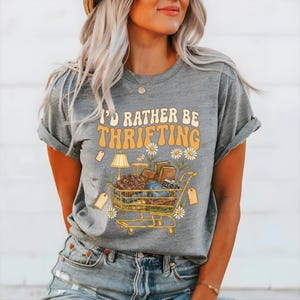 Op de afbeelding: Grijs t-shirt met de tekst "I'D RATHER BE THRIFTING" in oranje en geel. Een winkelwagenafbeelding is gevuld met tweedehands items, waaronder een lamp, boeken en een camera. Madeliefjes en prijskaartjes zijn ook te zien in het ontwerp.