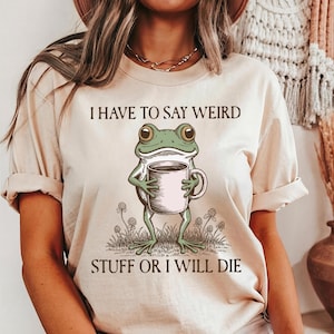 May include: Beige t-shirt with the text "I HAVE TO SAY WEIRD STUFF OR I WILL DIE." The shirt features a cartoon frog holding a coffee mug. The frog is green with a white belly and is standing in a patch of grass with clovers.