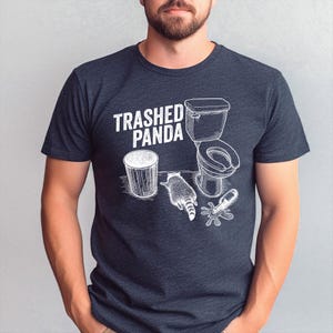 May include: Dark gray t-shirt with a white graphic of a raccoon, toilet, trash can, and spilled bottle, with the text "Trashed Panda". The design is a humorous illustration.