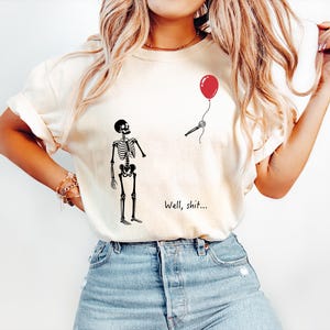Pode incluir: Uma t-shirt de cor creme com um gr&aacute;fico preto de um esqueleto de p&eacute; a olhar para uma m&atilde;o esquel&eacute;tica separada que segura um bal&atilde;o vermelho. O texto "Well, shit..." est&aacute; impresso a preto por baixo do esqueleto. A t-shirt tem mangas curtas.