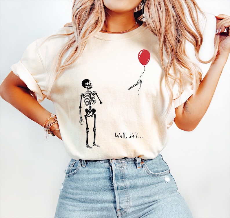 Pode incluir: Uma t-shirt de cor creme com um gr&aacute;fico preto de um esqueleto de p&eacute; a olhar para uma m&atilde;o esquel&eacute;tica separada que segura um bal&atilde;o vermelho. O texto "Well, shit..." est&aacute; impresso a preto por baixo do esqueleto. A t-shirt tem mangas curtas.