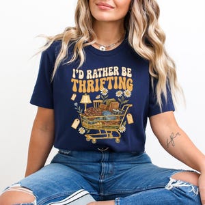 Op de afbeelding: Marineblauwe t-shirt met de tekst "I'd Rather Be Thrifting" in retrostijl. De afbeelding toont een winkelwagentje vol vintage items, labels en bloemaccenten. Een casual, leuk t-shirt.