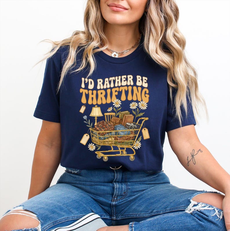 Op de afbeelding: Marineblauwe t-shirt met de tekst "I'd Rather Be Thrifting" in retrostijl. De afbeelding toont een winkelwagentje vol vintage items, labels en bloemaccenten. Een casual, leuk t-shirt.