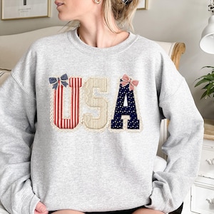 May include: A light grey sweatshirt featuring the word "USA" in a patriotic design. The letters are adorned with red and white stripes, beige lace, and blue with white stars. Each letter is embellished with a decorative bow.