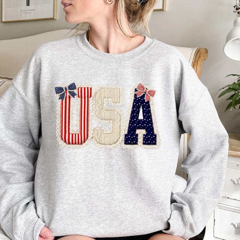 May include: A light grey sweatshirt featuring the word "USA" in a patriotic design. The letters are adorned with red and white stripes, beige lace, and blue with white stars. Each letter is embellished with a decorative bow.
