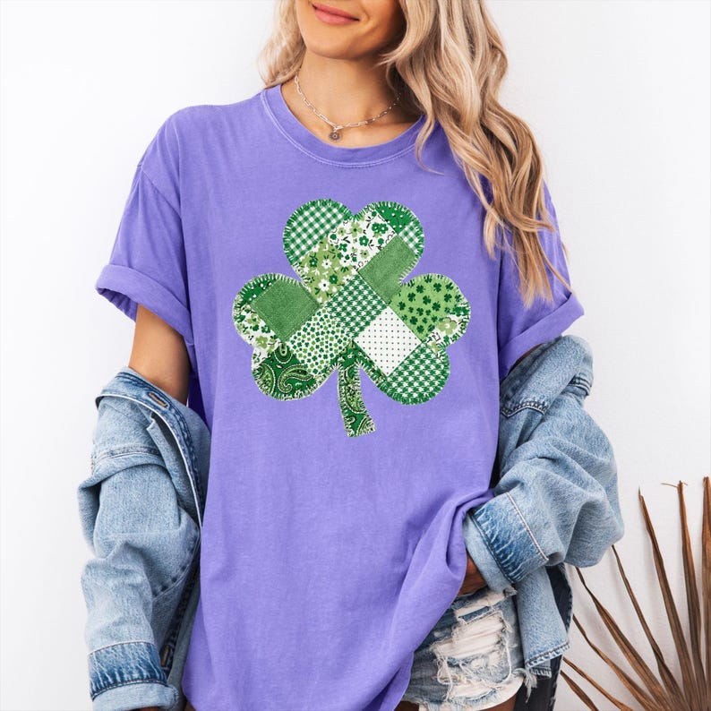 May include: A lilac-colored t-shirt featuring a patchwork shamrock design. The shamrock is composed of various green patterns, including gingham, floral, and paisley. The shirt has short sleeves and a relaxed fit, suitable for casual wear.