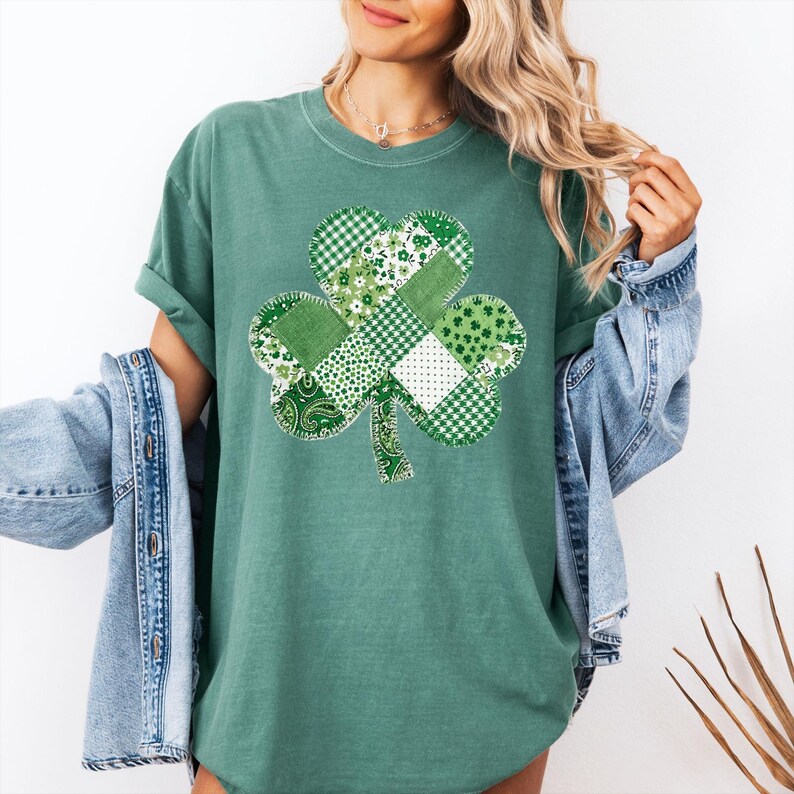 May include: Green t-shirt featuring a patchwork shamrock design. The shamrock is composed of various green patterns, including gingham, floral, and paisley. The shirt has short sleeves and a relaxed fit, perfect for St. Patrick's Day.