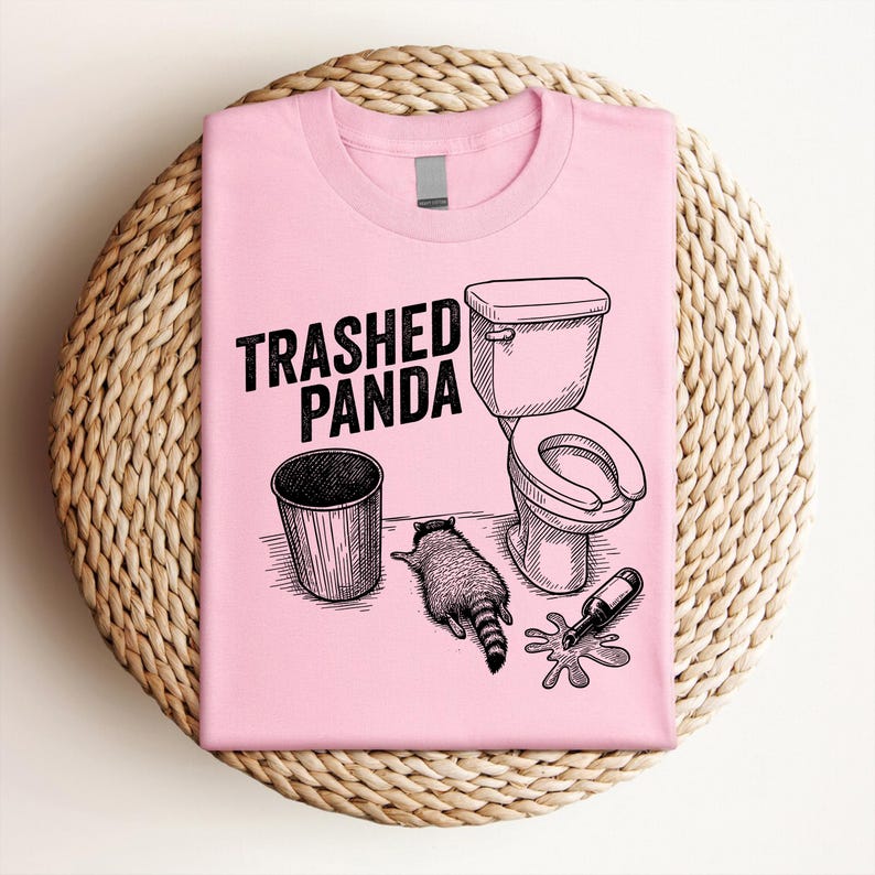 May include: A light pink t-shirt with the words "Trashed Panda" above a graphic of a trash can, toilet, raccoon, and spilled bottle. The design is in black ink.