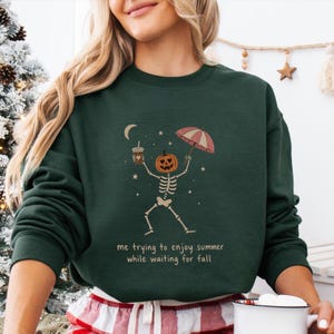May include: A dark green crewneck sweatshirt featuring a graphic of a skeleton with a pumpkin head holding a coffee cup and an umbrella. The text reads, "me trying to enjoy summer while waiting for fall," surrounded by small stars and a crescent moon.