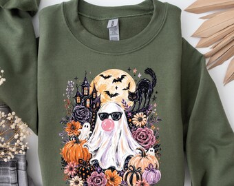 Peut inclure: Un sweat-shirt à col rond vert olive avec une impression graphique représentant un fantôme blanc portant des lunettes de soleil noires et faisant une bulle rose. Le motif inclut un manoir hanté, une pleine lune, des chauves-souris, un chat noir, des citrouilles et des fleurs colorées.
