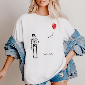 Pode incluir: Uma t-shirt branca com um gr&aacute;fico preto de um esqueleto de p&eacute; a olhar para uma m&atilde;o esquel&eacute;tica separada que segura um bal&atilde;o vermelho. O texto "Well, shit..." est&aacute; impresso a preto por baixo do esqueleto. A t-shirt &eacute; usada com um blus&atilde;o de ganga azul.