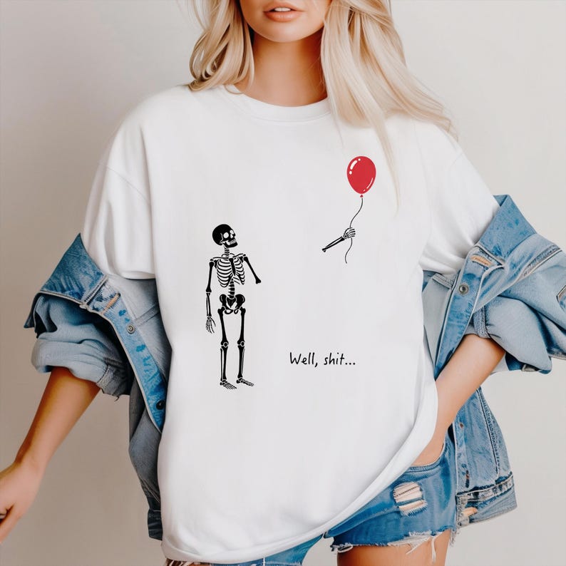 Pode incluir: Uma t-shirt branca com um gr&aacute;fico preto de um esqueleto de p&eacute; a olhar para uma m&atilde;o esquel&eacute;tica separada que segura um bal&atilde;o vermelho. O texto "Well, shit..." est&aacute; impresso a preto por baixo do esqueleto. A t-shirt &eacute; usada com um blus&atilde;o de ganga azul.