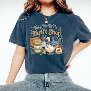 May include: A navy blue t-shirt with the text "Take Me To The Thrift Shop" in a floral design. The graphic includes a bread box, bowls, a duck, a chicken, and a mushroom.