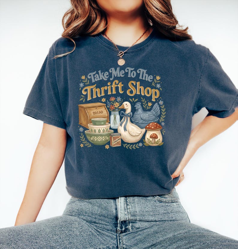 May include: A navy blue t-shirt with the text "Take Me To The Thrift Shop" in a floral design. The graphic includes a bread box, bowls, a duck, a chicken, and a mushroom.