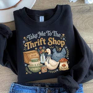 May include: Black sweatshirt with the text "Take Me To The Thrift Shop" in a vintage style. The graphic includes illustrations of a bread box, bowls, a duck, a chicken, and a mushroom. The sweatshirt is paired with blue jeans and a white hat.
