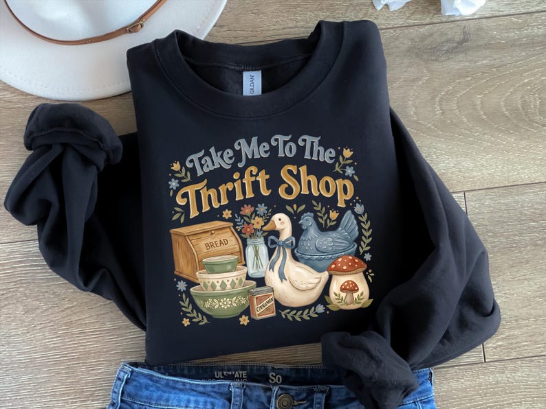 May include: Black sweatshirt with the text "Take Me To The Thrift Shop" in a vintage style. The graphic includes illustrations of a bread box, bowls, a duck, a chicken, and a mushroom. The sweatshirt is paired with blue jeans and a white hat.