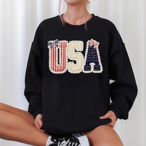 May include: Black sweatshirt featuring the word "USA" in a patriotic design. The letters are decorated with red and white stripes, blue polka dots, and a cream-coloured background, with decorative bows and a scalloped edge.