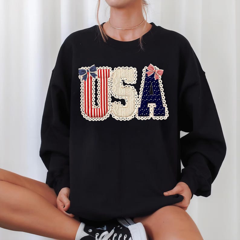 May include: Black sweatshirt featuring the word "USA" in a patriotic design. The letters are decorated with red and white stripes, blue polka dots, and a cream-coloured background, with decorative bows and a scalloped edge.