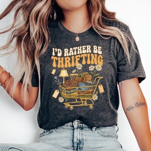 Op de afbeelding: Donkergrijs t-shirt met de tekst "I'd Rather Be Thrifting" in een retro lettertype. De afbeelding toont een winkelwagen vol vintage items, madeliefjes en een lamp. Het t-shirt wordt door een persoon gedragen.