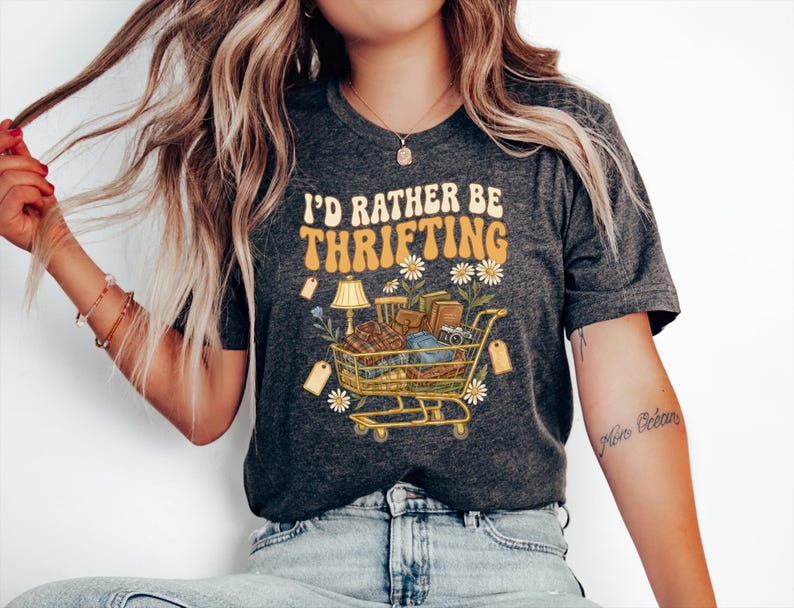 Op de afbeelding: Donkergrijs t-shirt met de tekst "I'd Rather Be Thrifting" in een retro lettertype. De afbeelding toont een winkelwagen vol vintage items, madeliefjes en een lamp. Het t-shirt wordt door een persoon gedragen.