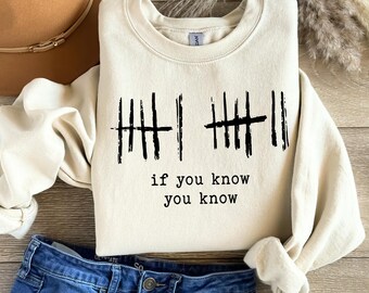 May include: Cream-colored sweatshirt with black tally marks and the phrase "if you know you know" printed in black. The sweatshirt is paired with blue denim jeans and a brown hat.