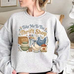 May include: A light gray sweatshirt with a graphic that says "Take Me To The Thrift Shop." The graphic includes a duck with a blue bow, a blue chicken, a mushroom, and other vintage-style illustrations. The sweatshirt is a comfortable, casual style.
