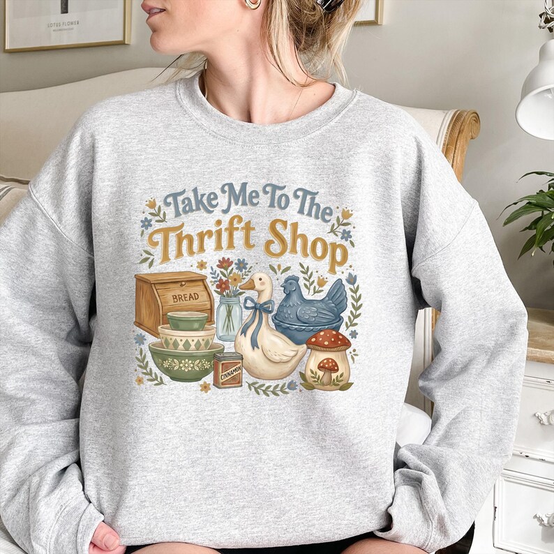 May include: A light gray sweatshirt with a graphic that says "Take Me To The Thrift Shop." The graphic includes a duck with a blue bow, a blue chicken, a mushroom, and other vintage-style illustrations. The sweatshirt is a comfortable, casual style.