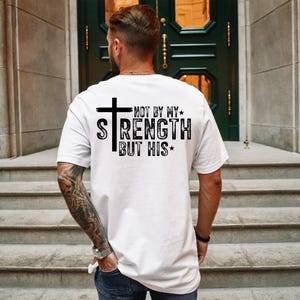 May include: White t-shirt with the text "NOT BY MY STRENGTH BUT HIS" in black, featuring a cross graphic. The shirt is a classic crew neck style.