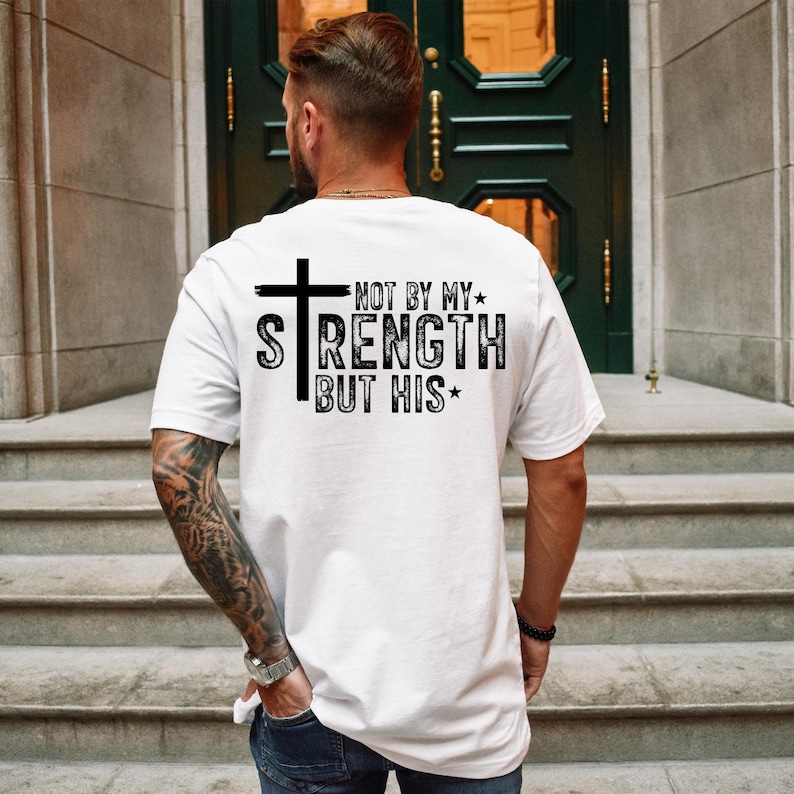 May include: White t-shirt with the text "NOT BY MY STRENGTH BUT HIS" in black, featuring a cross graphic. The shirt is a classic crew neck style.