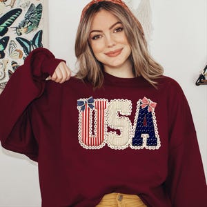 May include: Burgundy sweatshirt featuring the word "USA" in a patriotic design. The letters are decorated with red and white stripes, a beige textured "S", and blue stars. Each letter is outlined with white lace and topped with a bow.