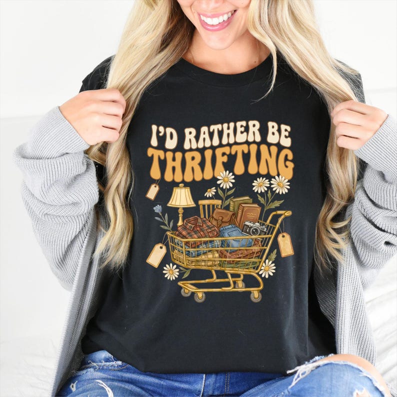 Op de afbeelding: Zwart t-shirt met de tekst "I'D RATHER BE THRIFTING" in oranje en wit. De afbeelding toont een winkelwagentje vol vintage items, waaronder een lamp, boeken en een camera, omringd door madeliefjes en prijskaartjes.