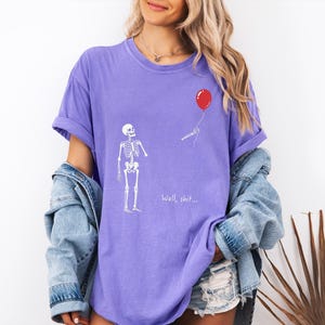 Pode incluir: Uma t-shirt roxa com um gr&aacute;fico branco de um esqueleto de p&eacute; ao lado de um bal&atilde;o vermelho que flutua para longe. O esqueleto estende a m&atilde;o em dire&ccedil;&atilde;o ao bal&atilde;o e o texto "Well, shit..." est&aacute; impresso em branco abaixo do esqueleto.