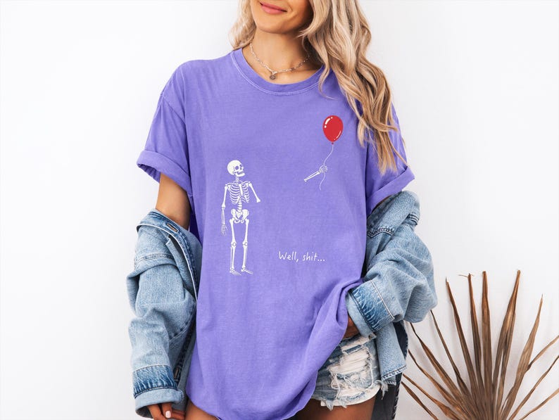 Pode incluir: Uma t-shirt roxa com um gr&aacute;fico branco de um esqueleto de p&eacute; ao lado de um bal&atilde;o vermelho que flutua para longe. O esqueleto estende a m&atilde;o em dire&ccedil;&atilde;o ao bal&atilde;o e o texto "Well, shit..." est&aacute; impresso em branco abaixo do esqueleto.