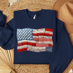 May include: Navy blue sweatshirt featuring a patchwork American flag design. The flag incorporates blue denim with white stars, and red and white patterned fabric stripes. The sweatshirt has a crew neck and long sleeves.