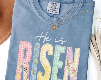Op de afbeelding: Een blauw Comfort Colors T-shirt met de tekst "He is Risen" in kleurrijke letters in patchwork-stijl. De letters zijn versierd met bloemen- en patroonstoffen. Een gouden ketting met een hanger ligt op het shirt.