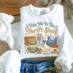 May include: White sweatshirt with the text "Take Me To The Thrift Shop" and a graphic of a duck, chicken, bread box, bowls, and a mushroom. The sweatshirt is paired with blue jeans and white sneakers.