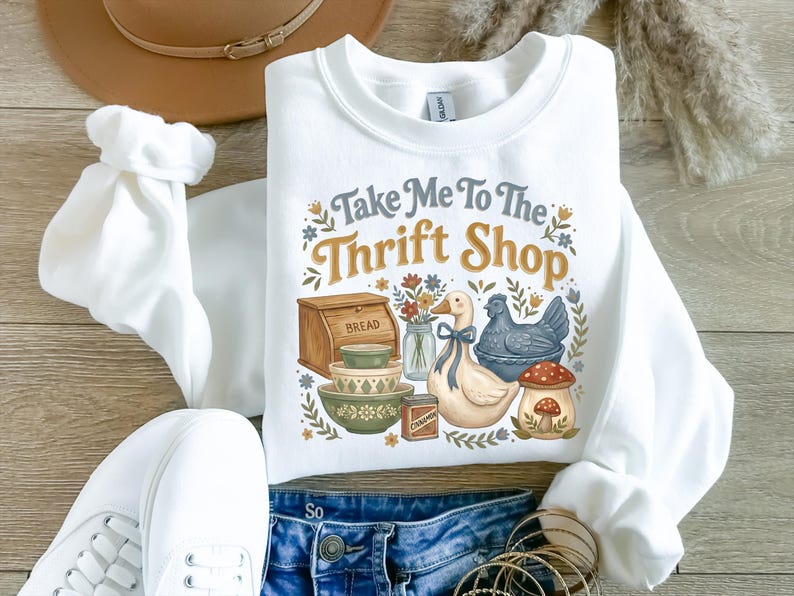 May include: White sweatshirt with the text "Take Me To The Thrift Shop" and a graphic of a duck, chicken, bread box, bowls, and a mushroom. The sweatshirt is paired with blue jeans and white sneakers.